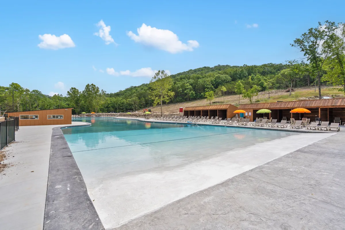 An inviting outdoor pool scene complete with colorful umbrellas, a wooden pavilion, and ample lounge chairs. The pool is surrounded by lush greenery, making it an ideal spot for swimming, sunbathing, and social gatherings in the warmer months.