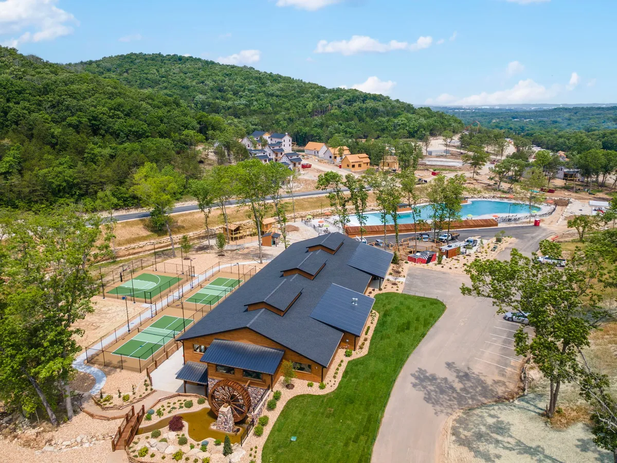 A broad view of the main building surrounded by lush greenery, showcasing a sparkling swimming pool and tennis courts nestled in nature. The image conveys a sense of community and shared outdoor amenities available within the resort.