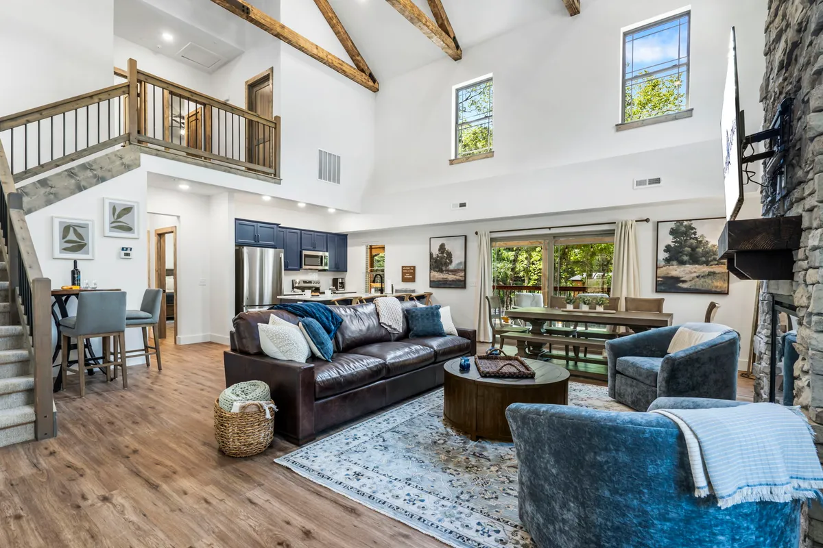 This image captures the open-concept gathering space with a large leather sofa, a stone fireplace as a central focal point, a spacious dining table with chairs, and expansive windows that welcome natural light. The scene feels both connected and relaxed, reflecting the heart of the home.