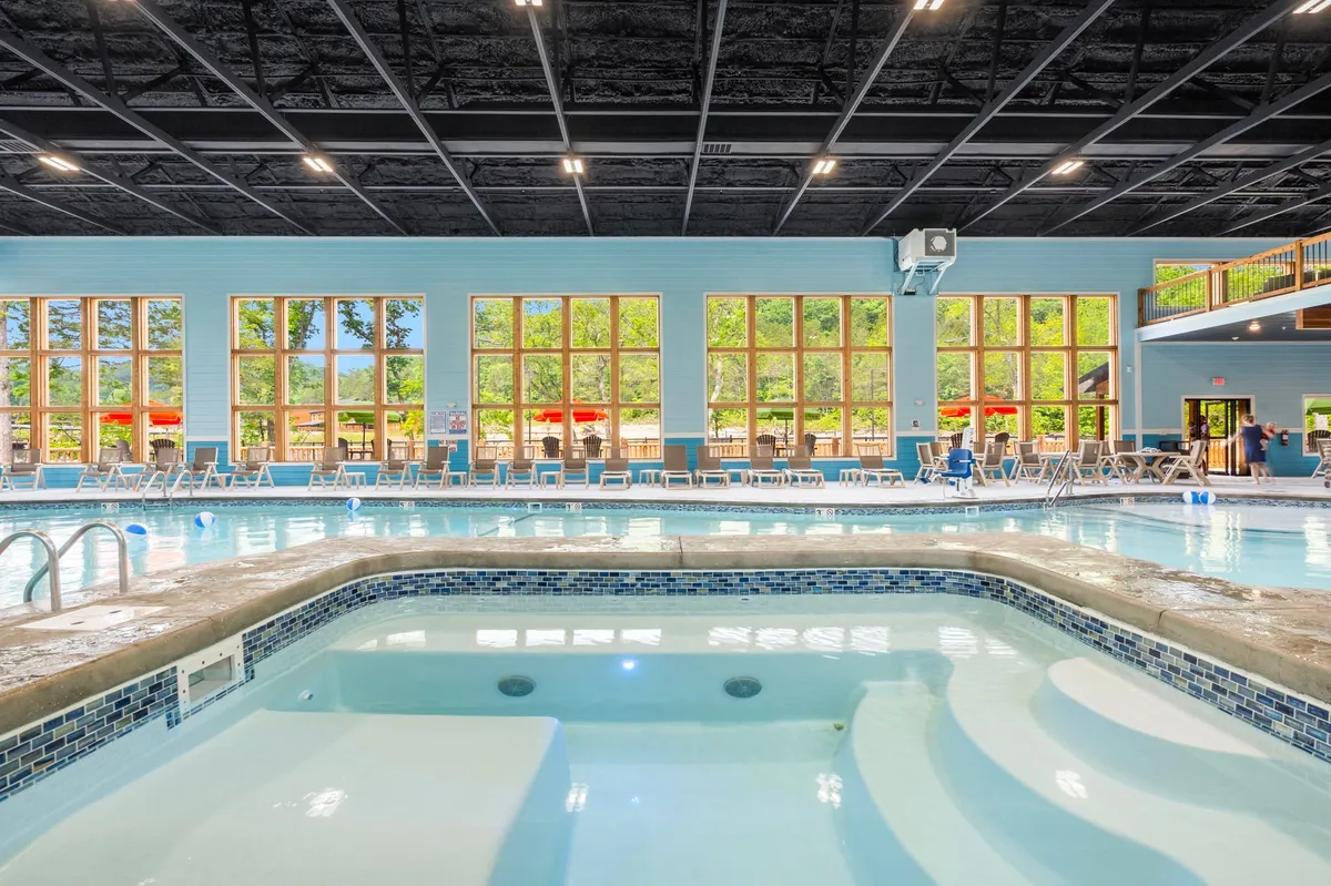 A bright, spacious indoor pool area featuring a large swimming pool and hot tub. Ample natural light pours in through expansive windows, while poolside seating invites guests to relax. The atmosphere is cheerful, ideal for swimming, relaxing in the hot tub, or socializing with friends and family.