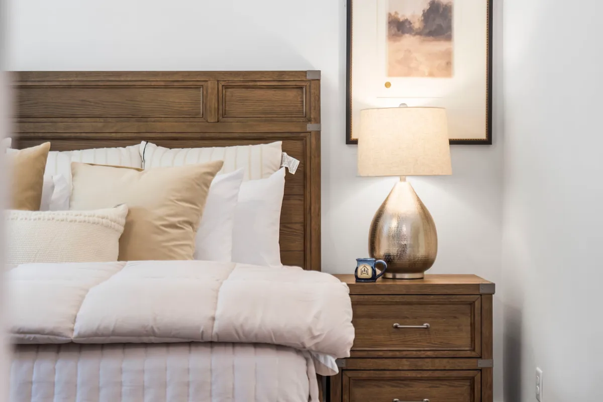 A serene bedroom featuring a bed with plush bedding, a golden lamp on a nightstand, and framed artwork on the wall. Natural light filters gently through the window, giving the room a cozy and peaceful feel.