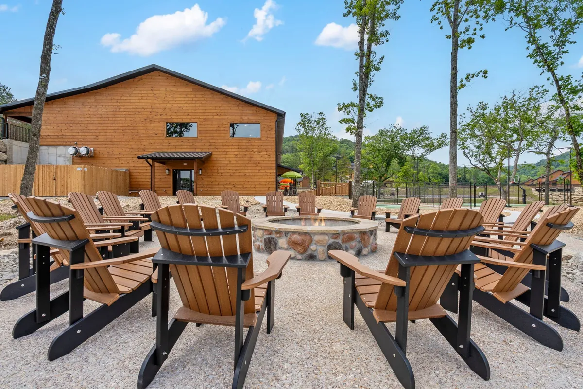 An inviting exterior communal space with Adirondack chairs arranged around a fire pit beside a wooden cabin. The scene suggests storytelling, casual barbecues, and laughter shared under the stars, all with wooded views in the background.
