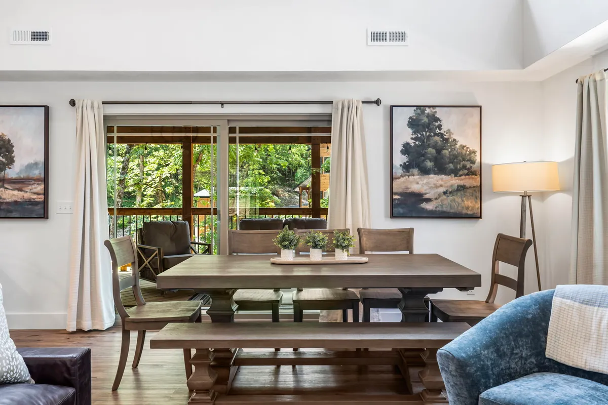 This image centers on a large dining table positioned near wide windows overlooking the woods, with distinctive artwork and a cozy lamp enhancing the atmosphere. It exudes warmth and encourages gatherings, whether for meals, games, or quiet moments.