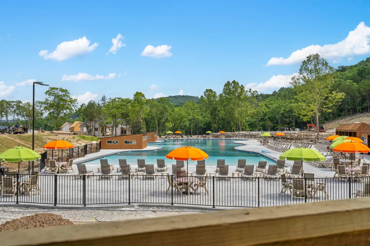 A vibrant outdoor pool scene surrounded by colorful umbrellas and lush green trees. Families can be imagined swimming, sunbathing, and relaxing together in this lively, sun-filled environment. The area offers plenty of space for parents and kids to enjoy warm afternoons.