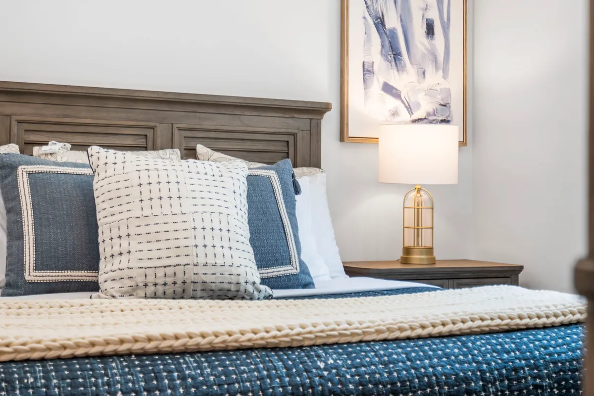 A cozy bedroom with a bed adorned with textured blankets and decorative pillows. A nightstand with a lamp and framed artwork above the bed add to the room's tranquil, inviting atmosphere.