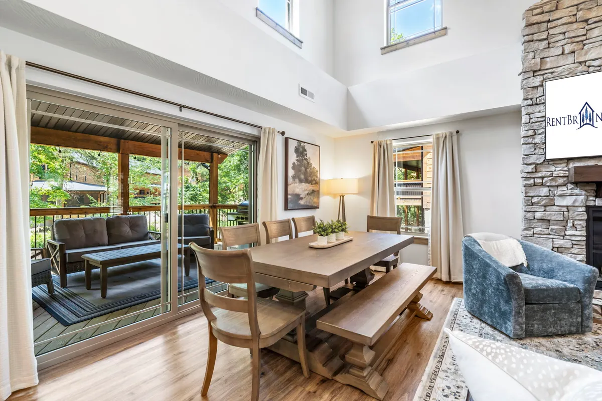 A wide view featuring a dining table set for a meal, a comfortable sofa, the glowing fireplace, and expansive windows with views of the outdoors. Decorative artwork and warm lighting reinforce a sense of welcome and comfort.