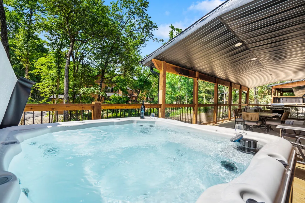 The image showcases the exterior of a beautiful lodge nestled in lush greenery, highlighting a private hot tub surrounded by nature and a comfortable outdoor seating area. The atmosphere is tranquil, inviting guests to relax, unwind, and enjoy the serene Ozark setting with friends or family.